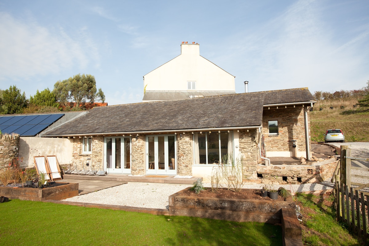 The exterior of The Piggery showcases a combination of natural stone and modern design, featuring large glass doors that allow light to flood in. A generously sized lawn area with gravel paths extends in front, while solar panels are discreetly positioned on the roof.