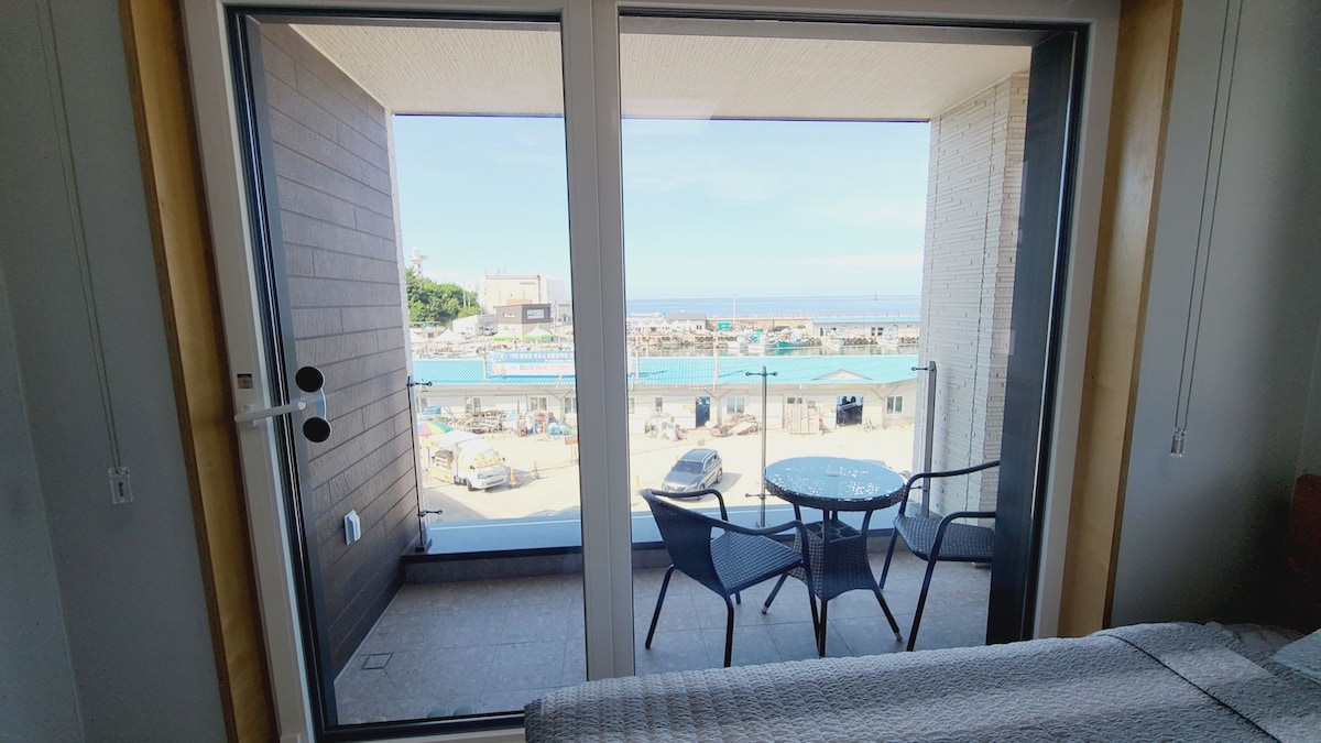 A bright view is captured through sliding glass doors, which open to a small balcony. Two chairs and a table are situated on the balcony, overlooking the bustling Aeyajin port and the expansive ocean. Natural light fills the room, enhancing the tranquil atmosphere.