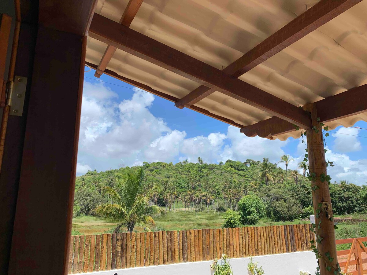A view from a covered porch reveals lush greenery, with palm trees and dense vegetation contrasting against a clear blue sky. The wooden railing frames the scenery, while a fenced area demarcates the property, enhancing the surroundings' natural appeal.
