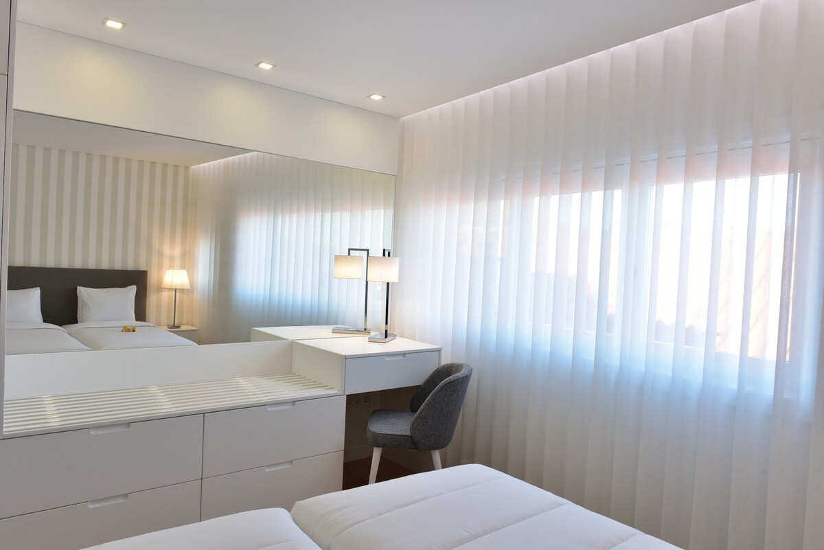 A well-lit bedroom features soft, neutral-toned walls with a large mirror reflecting light. A spacious bed is accompanied by a sleek bedside lamp and a modern desk with an ergonomic chair. Sheer curtains partially cover the window, allowing natural light to enhance the room's ambiance.