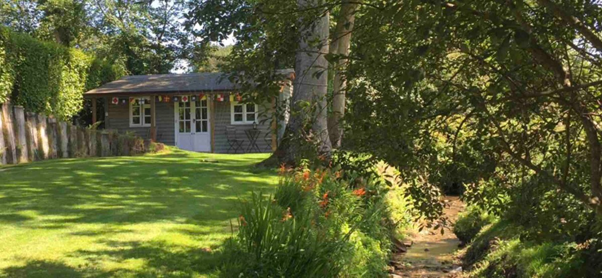 The studio cabin is nestled in a lush garden, surrounded by greenery and wildflowers. A small stream flows nearby, with the cabin's entrance facing the well-kept lawn. Lanterns hang subtly, adding charm to the tranquil setting.