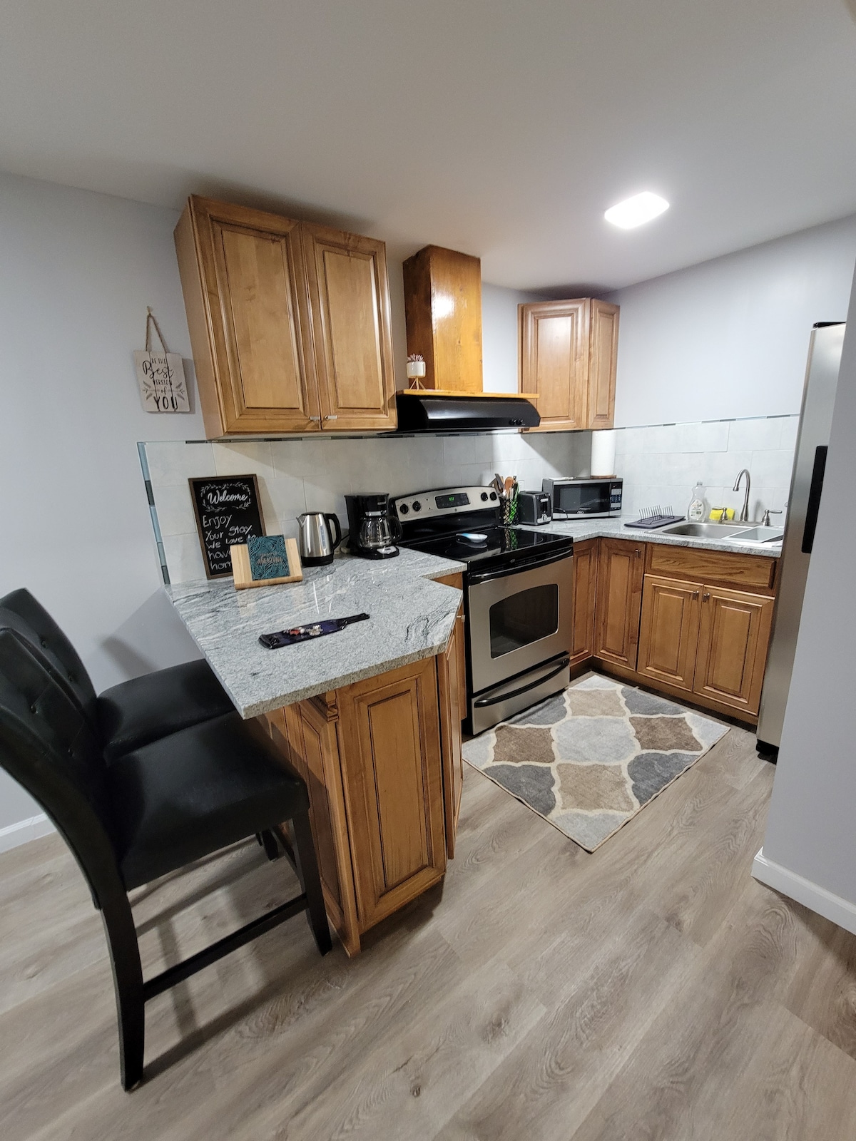A modern kitchen is displayed with wooden cabinets and granite countertops. Stainless steel appliances, including a stove and refrigerator, are present. A small dining area features two high-backed chairs beside a compact counter. A decorative rug adds texture to the space.