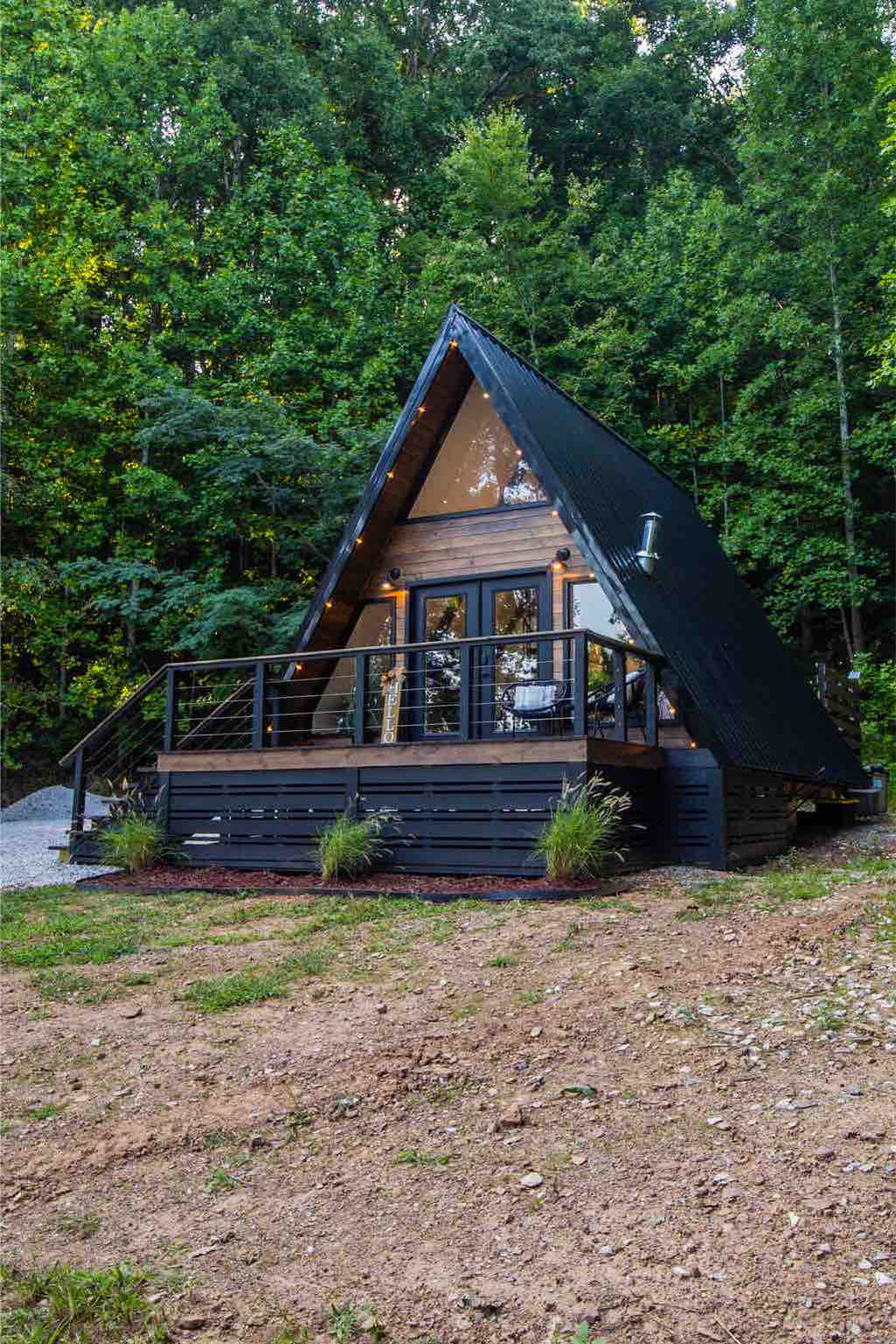 The exterior of a charming a-frame cabin is surrounded by lush green trees. A spacious wooden deck with black railings offers a welcoming entrance. Large windows provide glimpses of the interior, while gravel pathways lead to the cabin, framed by small plants and natural landscaping.