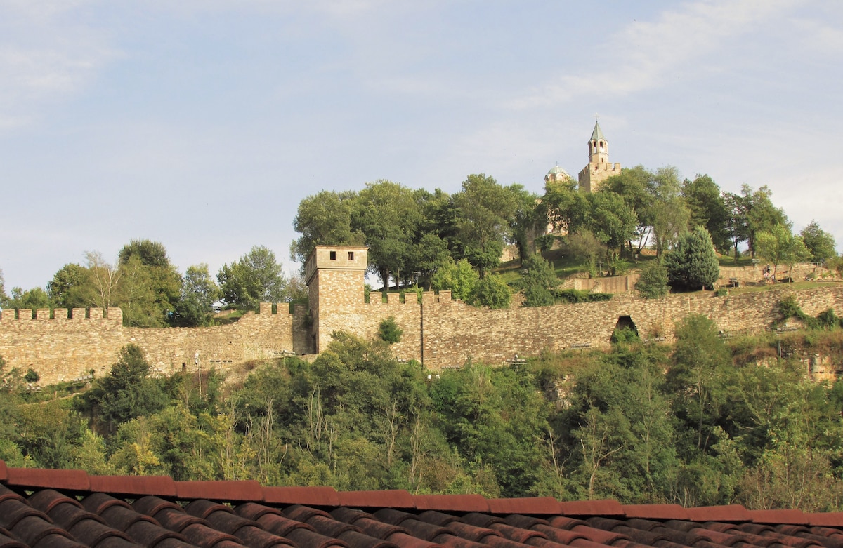 A panoramic view of a medieval fortress atop a hill, surrounded by lush greenery. The stone walls of the fortress are visible, along with a tower and a church-like structure featuring a steeple. The foreground displays a terracotta roof, blending with the natural landscape.