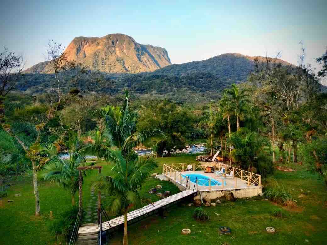 A scenic view reveals a vibrant outdoor area with a small pool surrounded by lush greenery. Towering mountains rise majestically in the background, and palm trees enhance the natural landscape. A wooden walkway leads to the pool area, inviting relaxation.