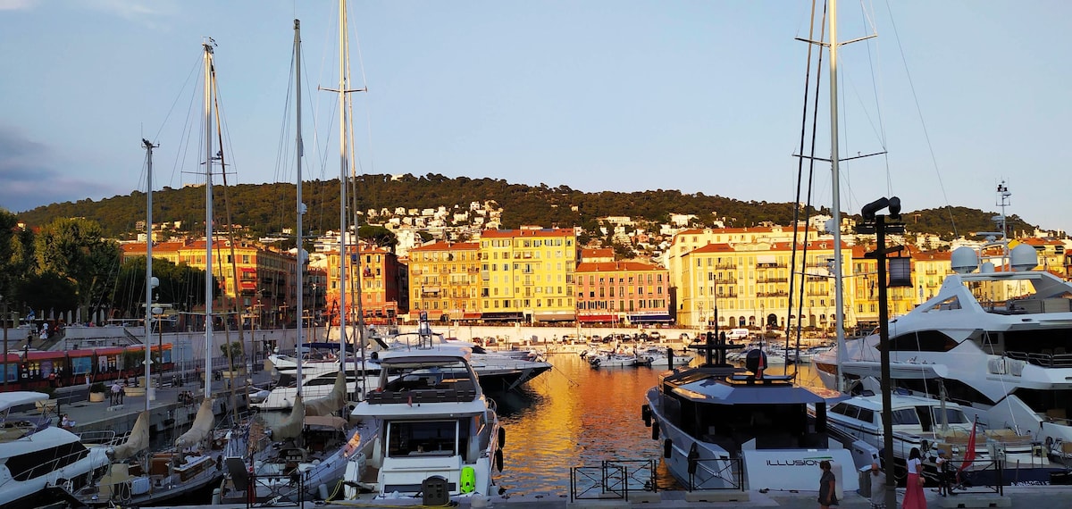 The image showcases a vibrant harbor with numerous boats docked near colorful buildings reflecting the warm light of sunset. A hillside is visible in the background, adorned with trees and residential areas, creating a scenic waterfront atmosphere.