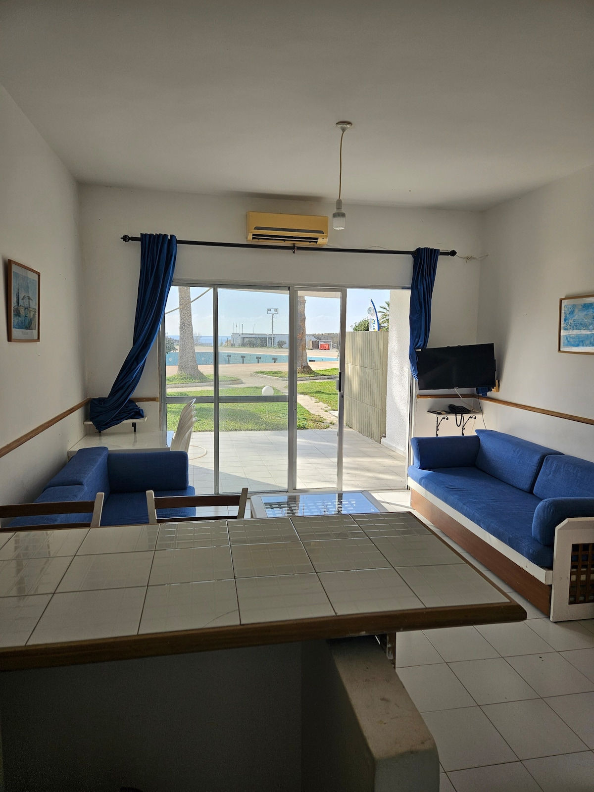 A light-filled living area features large glass doors that open to a view of the outdoors. Two blue sofas are positioned comfortably, with a small TV mounted on the wall. Soft, blue curtains frame the windows, adding a calming touch to the space.