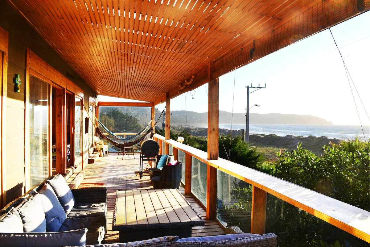 A spacious terrace is shown with wooden decking, featuring comfortable seating arrangements and a hammock. Glass railings provide unobstructed views of the ocean and surrounding natural scenery, with sunlight illuminating the area. The tranquil setting encourages relaxation and connection with nature.