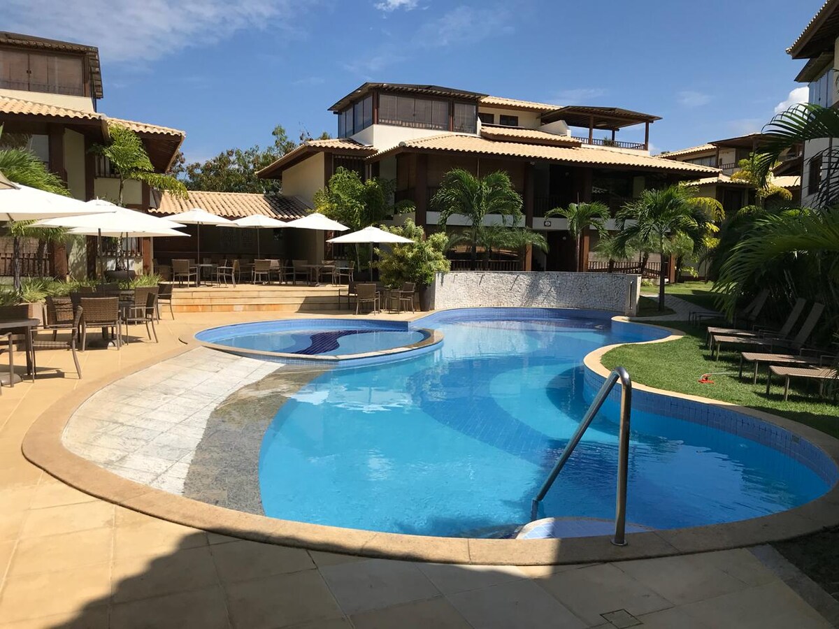 A large, inviting pool area is surrounded by lush greenery and palm trees. Umbrellas and outdoor dining tables are set along the poolside, while two circular hot tubs are integrated into the pool design. Sunlight reflects off the water, providing a serene atmosphere.