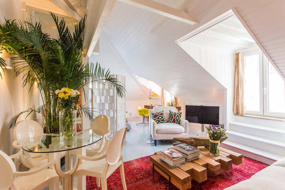 A bright and airy living area is shown, featuring a glass dining table surrounded by white chairs. An inviting sofa is positioned across from a television, and a coffee table made of wooden blocks holds a stack of magazines. Lush greenery adds a fresh touch.