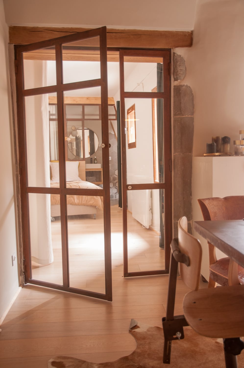 A wooden-framed door stands open, revealing a bright interior space. Light filters through the glass panes, highlighting the wooden flooring. In the background, a stylish bedroom can be seen, with a bed and circular mirror, adding depth to the image.