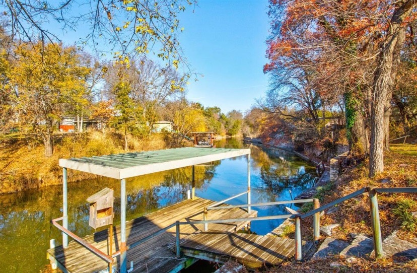 A wooden dock extends over calm waters, framed by trees displaying vibrant autumn foliage. Steps lead down to the dock, while reflections of the surrounding landscape are visible in the water, creating a serene outdoor environment.