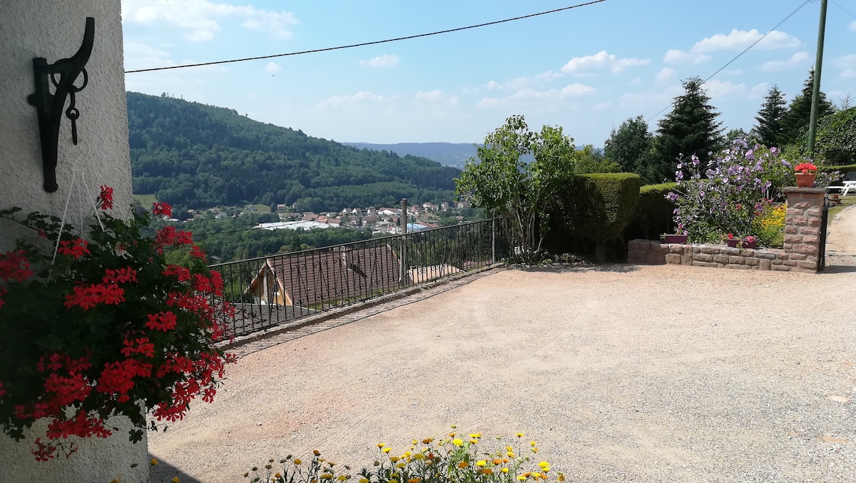 A gravel parking area is bordered by lush greenery and colorful flower beds, featuring vibrant red geraniums and yellow blooms. In the background, a panoramic view of the village and surrounding mountains is visible under a clear blue sky.