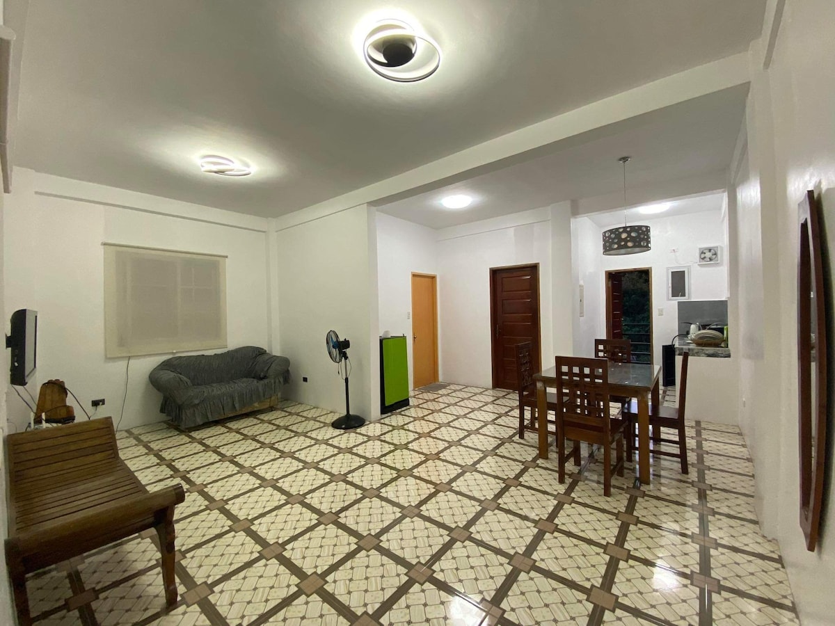 A spacious living area features a patterned tile floor, with a comfortable sofa placed against the wall. A wooden dining table with chairs occupies one side, while a green cabinet and a standing fan are visible. Natural light fills the room through a window.