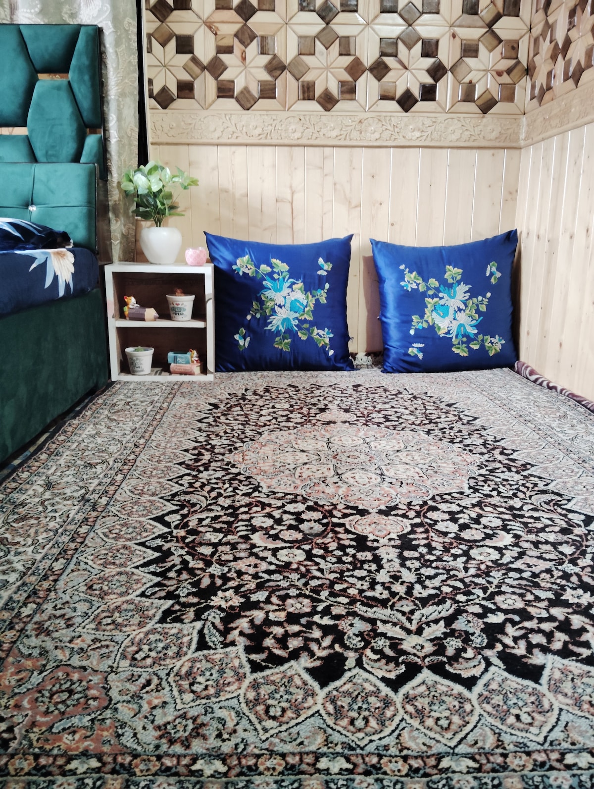 A cozy interior space features a detailed area rug with floral patterns in muted colors. Soft blue cushions with green designs are placed against a wooden backdrop. A small shelf holds potted plants and decorative items, creating a warm and inviting corner.