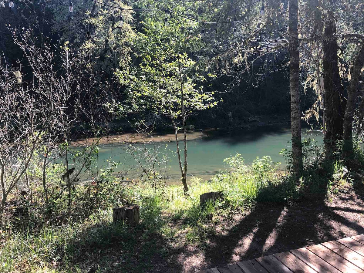The image captures a serene view of the river surrounded by lush greenery. Tall trees frame the scene, while sunlight filters through the leaves, illuminating the gentle water. A patch of grass and a wooden deck border the river, enhancing the tranquil atmosphere.