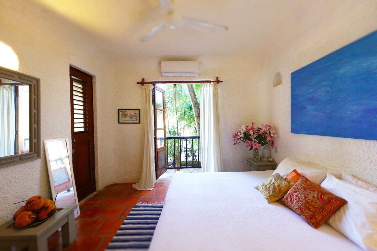 A bright bedroom features a spacious bed dressed in white linens, with colorful pillows adding contrast. An artwork in blue adorns the wall. Natural light enters through double doors that open to a small balcony surrounded by greenery, complemented by a decorative mirror and potted flowers.