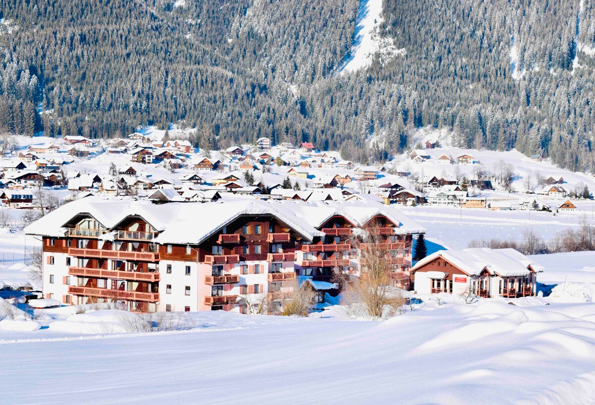 A winter scene depicts a collection of buildings blanketed in snow, nestled among gently sloping hills. The surrounding landscape includes a range of houses and trees, showcasing a serene alpine environment under a clear sky.