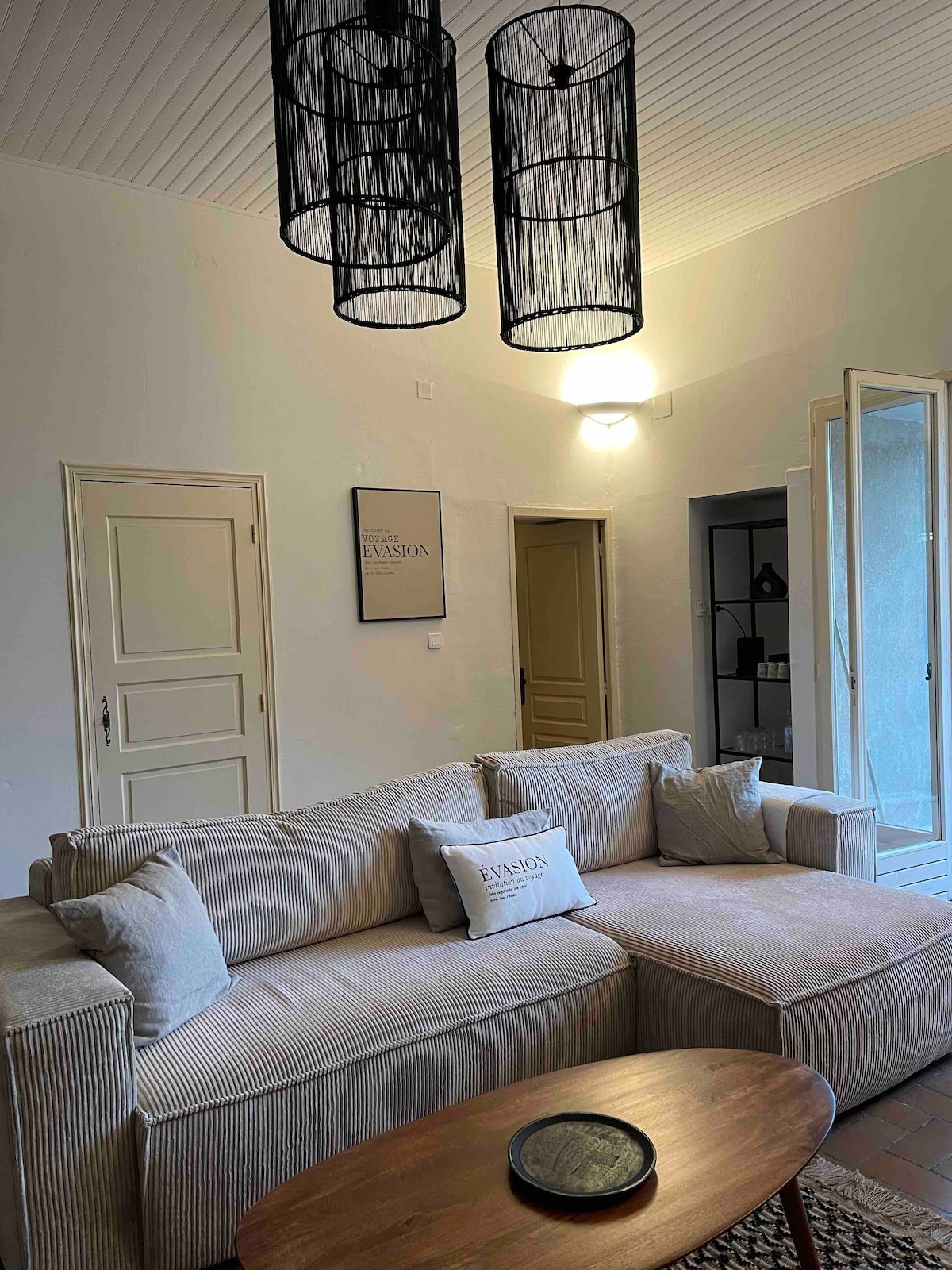 A spacious living area is featured, showcasing a comfortable sectional sofa adorned with decorative pillows. Soft lighting is provided by three pendant lamps hanging from the ceiling. Two doors lead to undefined areas, while a wooden coffee table rests in front of the sofa.