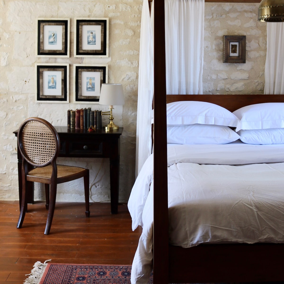 A spacious bedroom features a four-poster bed dressed in crisp white linens. A wooden desk with a brass lamp and a bookshelf sits beside the bed. Four framed artworks adorn the stone wall, while a woven chair adds a touch of comfort.