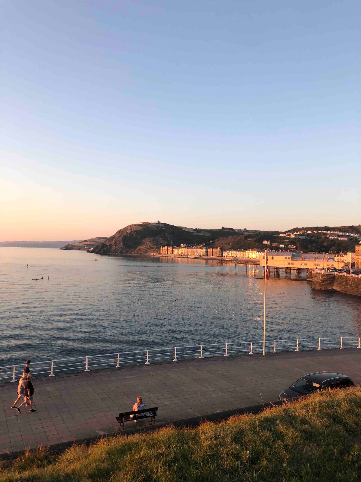 The coastline of Aberystwyth is captured during sunset, showcasing calm waters reflecting pastel hues. A waterfront promenade is visible with people strolling along the path. The backdrop features rolling hills and buildings that line the shore, creating a serene seaside atmosphere.