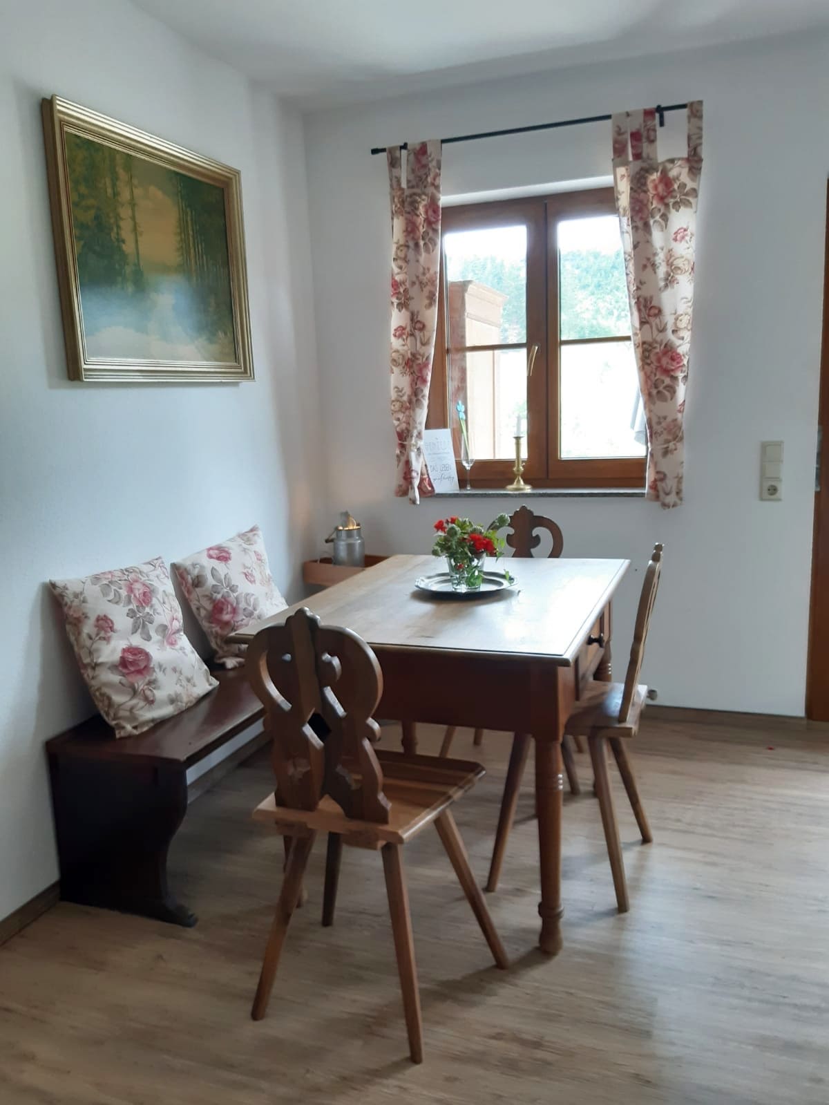 A dining area features a wooden table surrounded by three chairs. Floral-patterned cushions are placed on a wooden bench. A small vase with fresh flowers sits at the center of the table. Light enters through a large window, adorned with floral curtains.