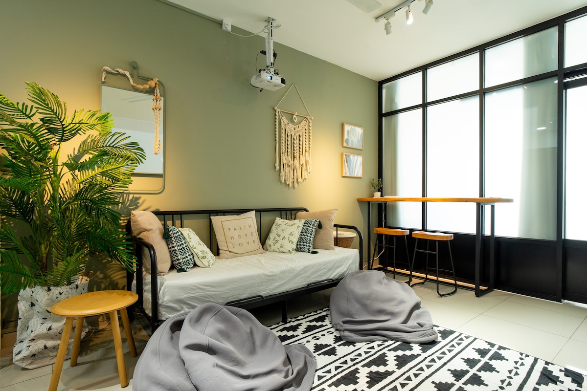 A bright living room is featured, showcasing a black sofa bed with decorative pillows. A wooden stool and a table are positioned near a large window, allowing natural light to enhance the space. A projector hangs from the ceiling, with a bohemian wall hanging and greenery adding to the decor.