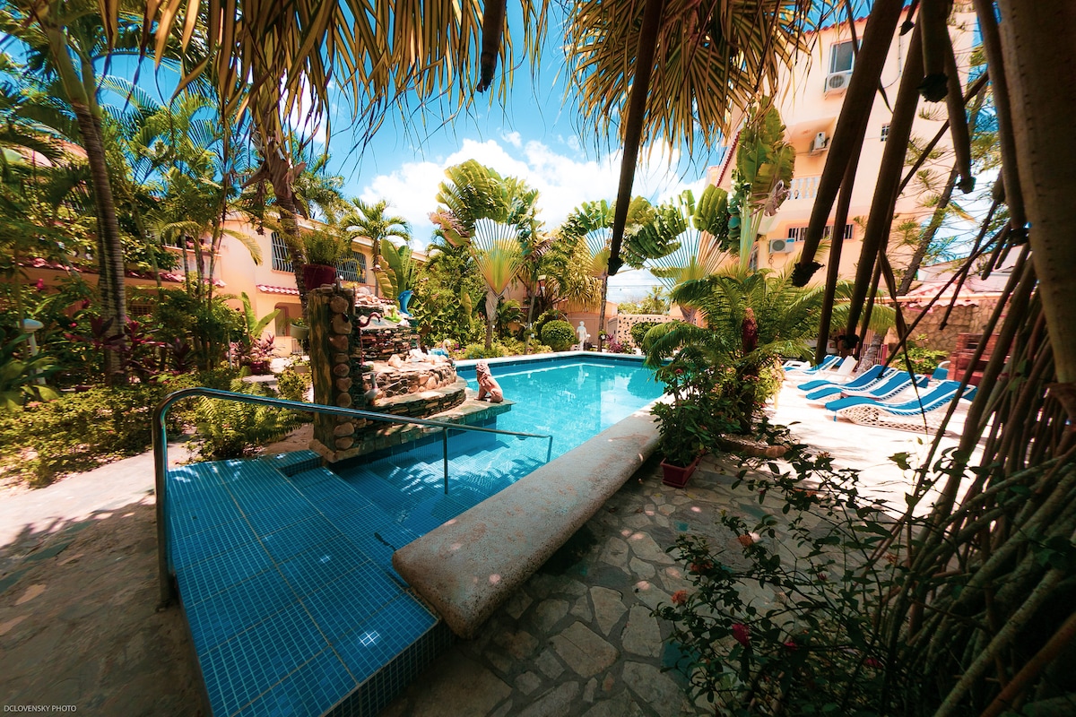 A shared outdoor pool is surrounded by lush tropical landscaping. Comfortable lounge chairs are positioned nearby, under the shade of palm trees. A stone fountain adds a tranquil element to the space, creating a serene retreat for relaxation.