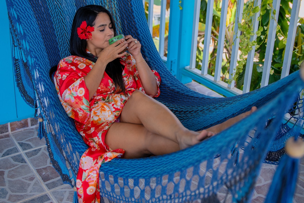 A vibrant blue hammock is positioned in a lush outdoor setting. A person relaxes comfortably, wearing a floral robe, while holding a drink. Surrounding greenery and bright blue railings enhance the tranquil atmosphere, creating an inviting space for leisure.