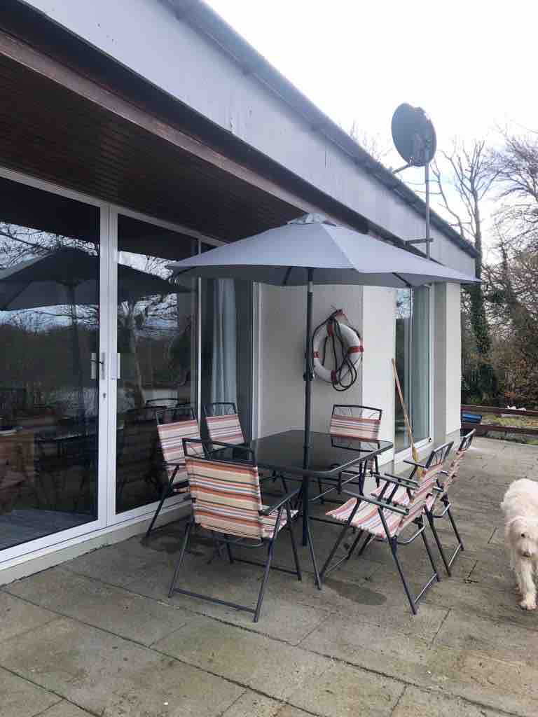 An outdoor dining area features a large table surrounded by six striped chairs, with a large umbrella providing shade. A life ring is mounted on the wall nearby, and lush greenery is visible in the background. The patio area is paved with gray tiles.
