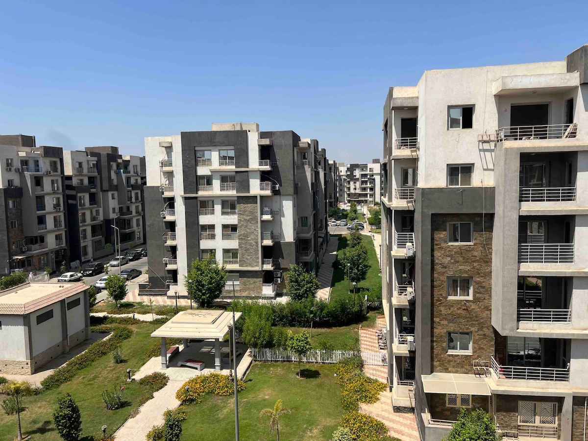 A view of a landscaped area features modern, multi-story buildings surrounded by green lawns and trees. Walkways meander through the grass, leading to shaded seating areas. Clear blue skies provide a bright backdrop to the contemporary architecture.