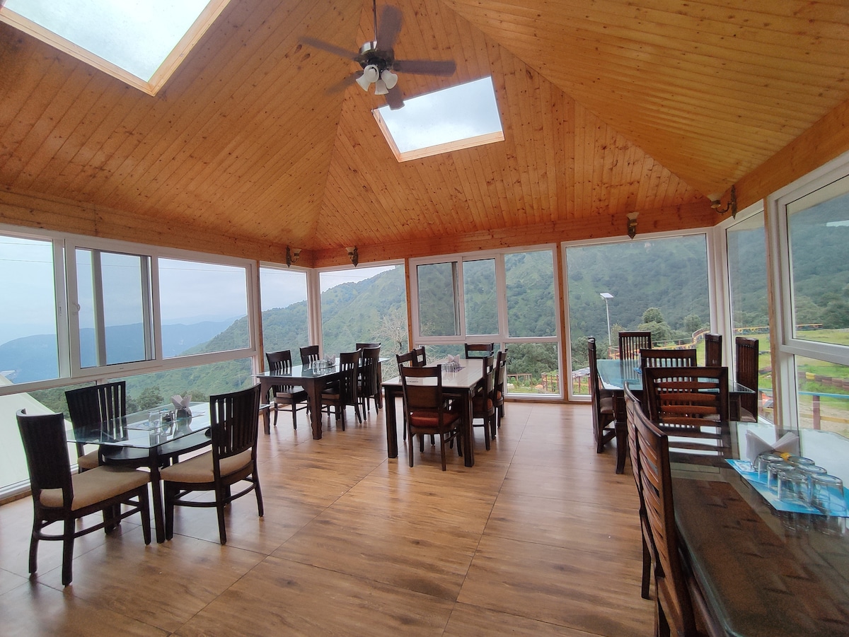A spacious dining area features large windows and skylights, allowing natural light to illuminate the wooden interior. Several tables and chairs are arranged to accommodate guests, while scenic views of the surrounding mountains can be seen through the windows.