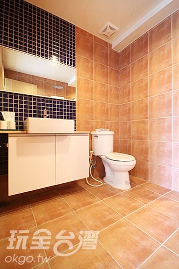 A modern bathroom features tiled walls in warm tones. A white sink is situated atop a minimalist cabinet below a mirror. A toilet is positioned beside the cabinet, with ample floor space observed for easy movement.