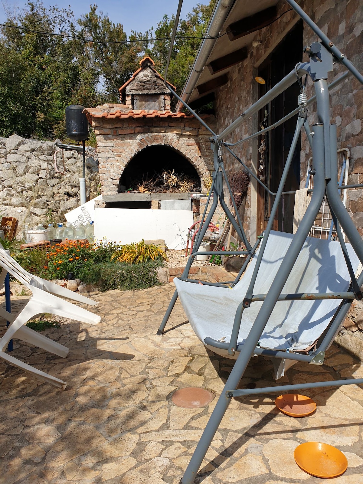 An outdoor space features a stone patio with a swing seat and two white lounge chairs. A traditional stone barbecue is visible in the background, surrounded by greenery and flowers. The area is well-maintained and offers a sense of privacy and calm.