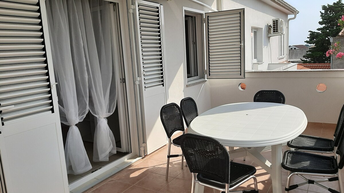 A terrace is shown, featuring a round white table surrounded by six black chairs. Two sliding doors with white curtains open to the outside, allowing natural light into the area. The tiles are light-colored, contributing to a bright and open feel.