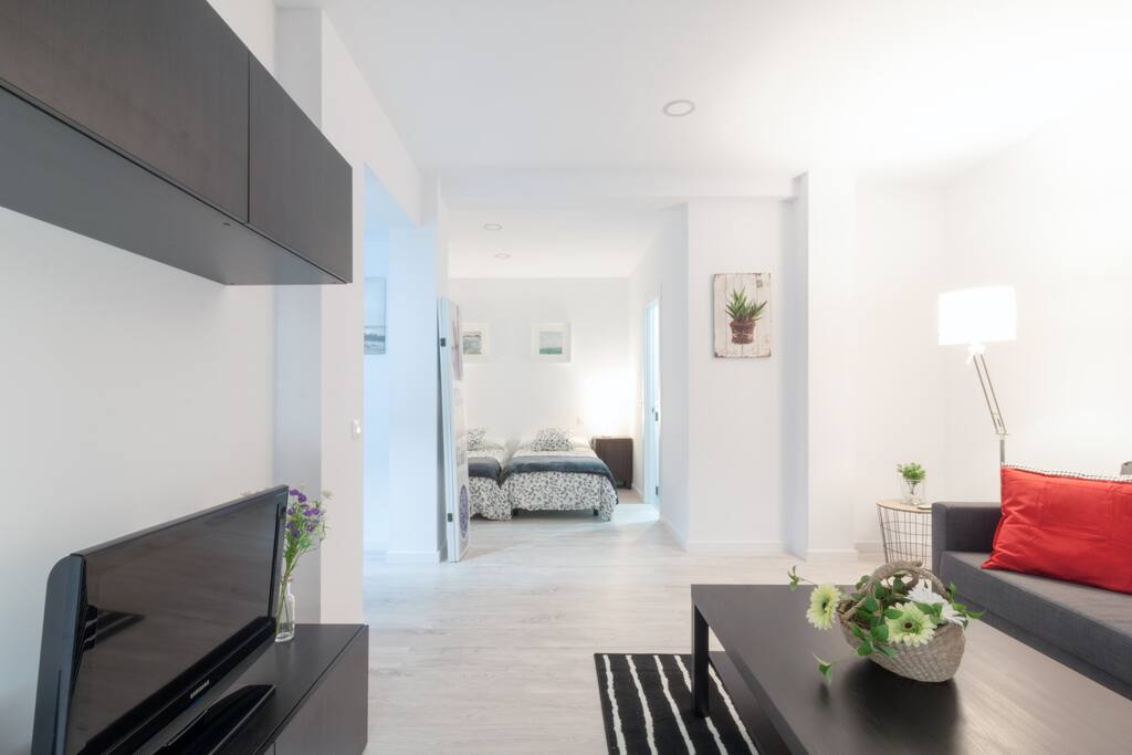Moderno y luminoso salón-comedor con sofá cama y TV de pantalla plana. Al fondo, se encuentra el dormitorio.