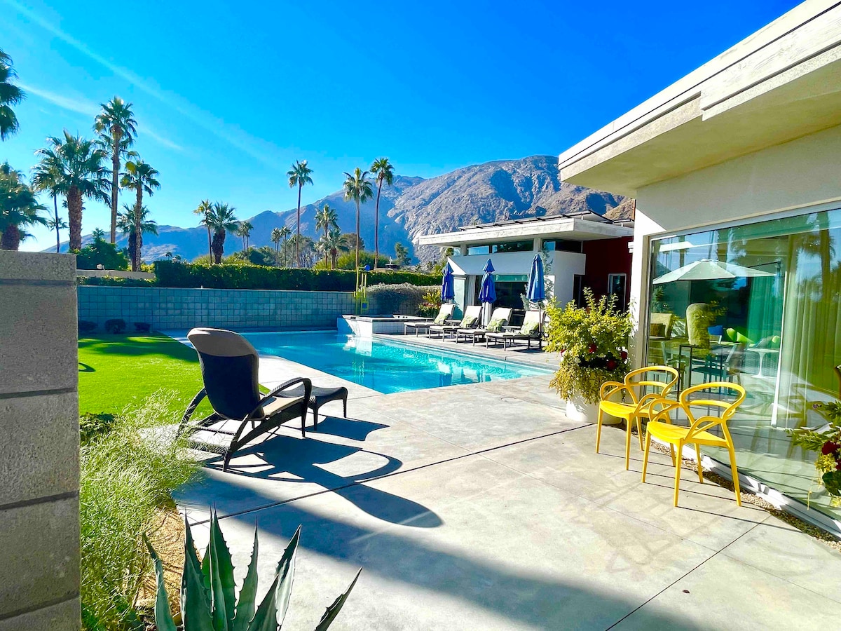 A spacious outdoor area features a large pool surrounded by lounge chairs and umbrellas. Lush green grass is present alongside palm trees and mountain views in the background. A pathway leads to the modern home's glass doors, inviting natural light into the interior.