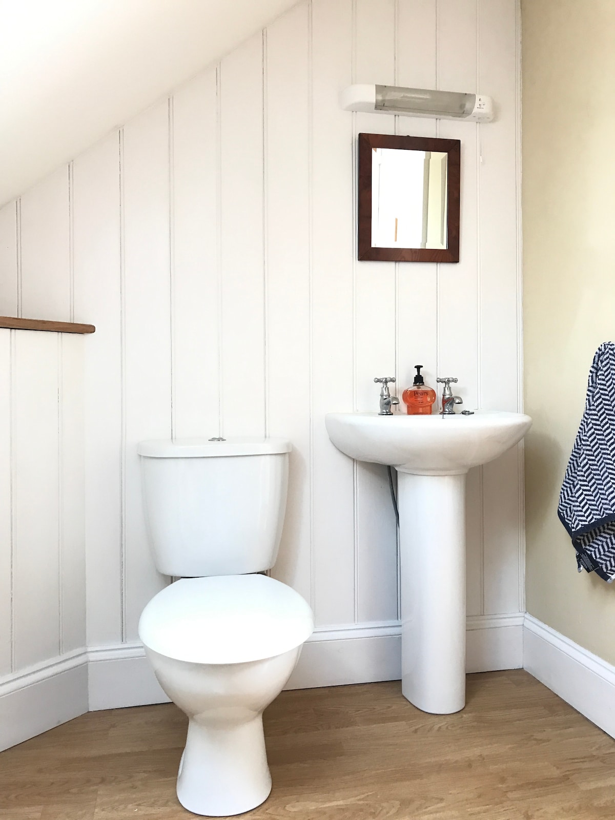 A bright bathroom features white wood paneling, creating a clean and fresh environment. A modern toilet and a pedestal sink with a small mirror above are present. Natural light enters from a nearby window, enhancing the uncluttered space.