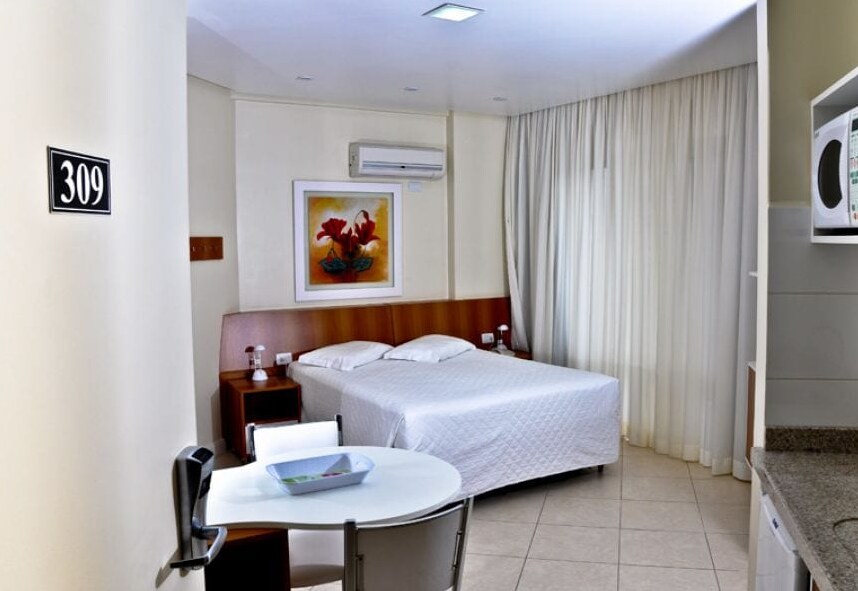 A well-lit room features a spacious bed with crisp white linens, paired with bedside tables and lamps. A dining area with a round table and chairs is present. The kitchenette includes a microwave, and large windows covered with light curtains allow for natural light.