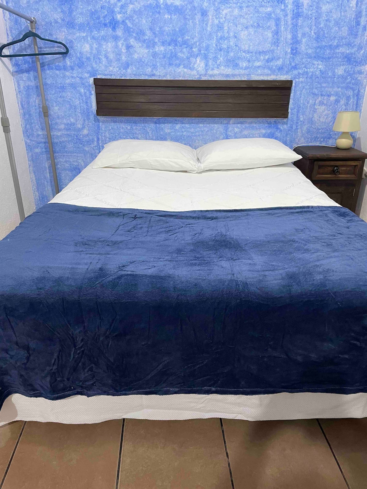 A simple bedroom features a spacious bed adorned with a navy blue blanket. Soft white bedding is complemented by a light-colored headboard. A small nightstand with a lamp is positioned beside the bed, and a clothing rack stands nearby against a textured blue wall.