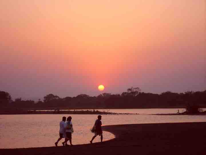 A serene riverside scene captures the sun setting beyond the horizon, casting warm hues of orange and pink across the sky. Silhouettes of two individuals are seen walking along the shore, with gentle ripples in the water reflecting the soft light.