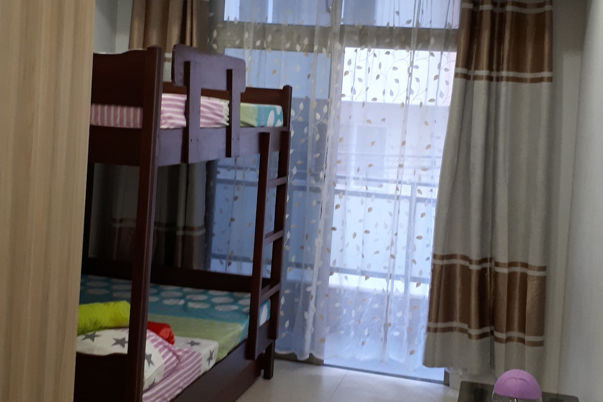 A cozy bedroom features a wooden bunk bed adorned with colorful bedding. Natural light filters through sheer curtains, illuminating the room. A small round object is visible on the floor, adding a playful touch to the space.