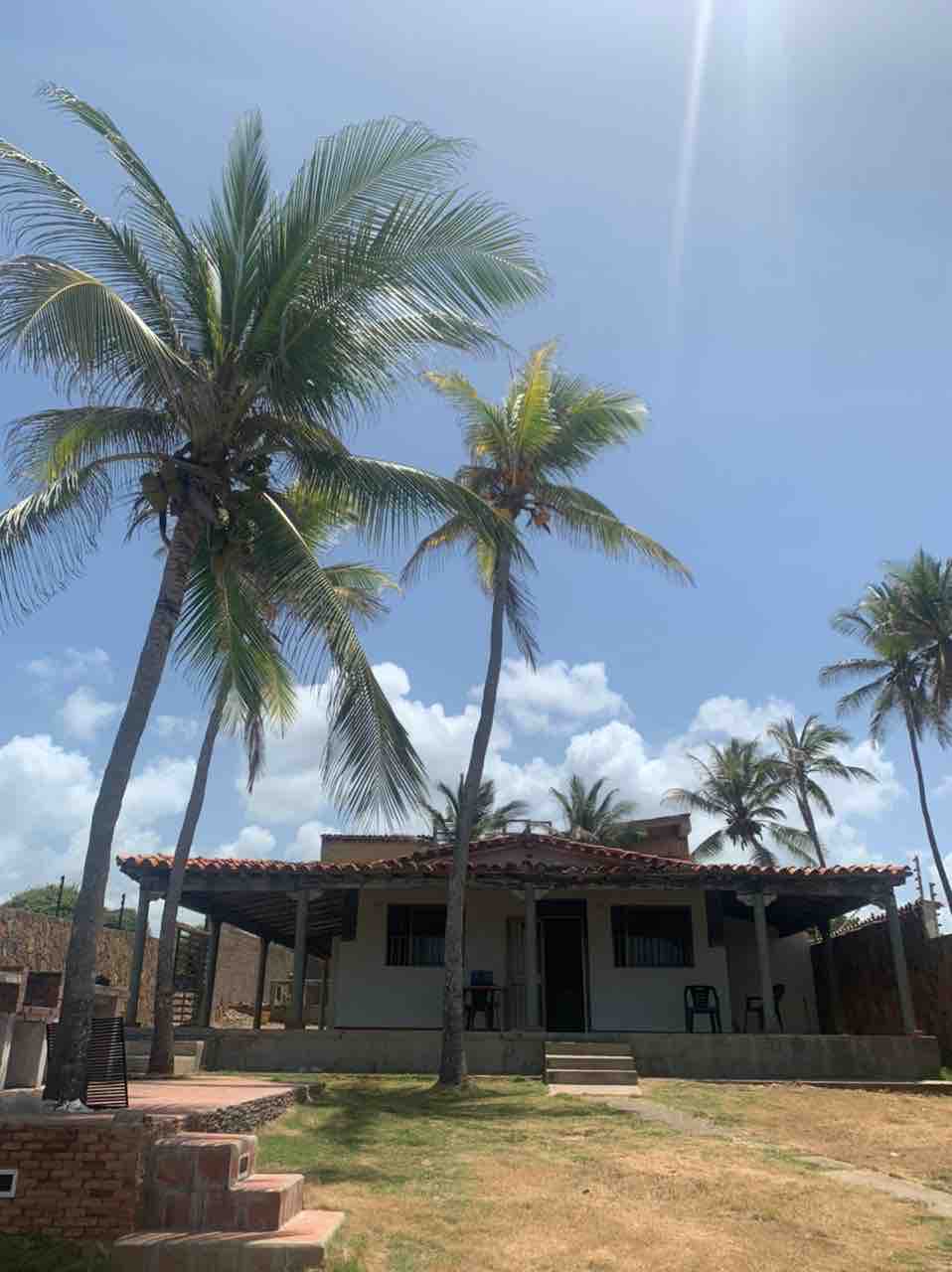 A charming beach house is showcased beneath a clear blue sky, surrounded by tall palm trees. The property features a covered porch that offers shaded seating, with steps leading to a grassy area in front. A rustic stone structure is partially visible on the left.