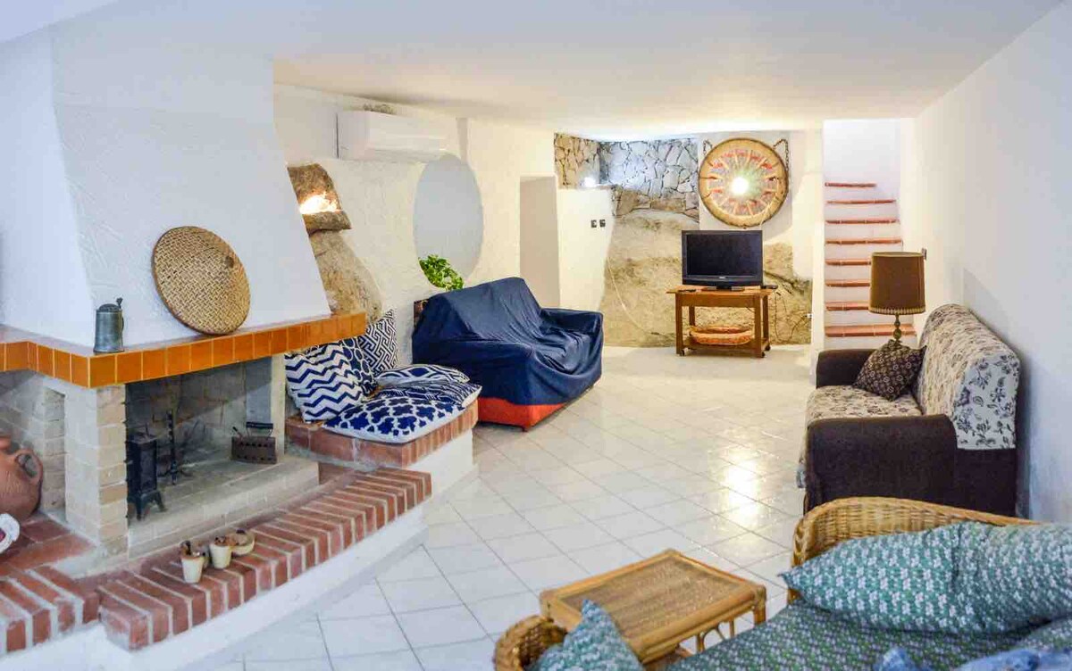 A living area features a fireplace surrounded by comfortable seating, including a patterned couch and a rattan chair. A TV is positioned near a patterned wall, while light is provided by wall fixtures. Stairs lead to an upper level, enhancing the spacious feel.