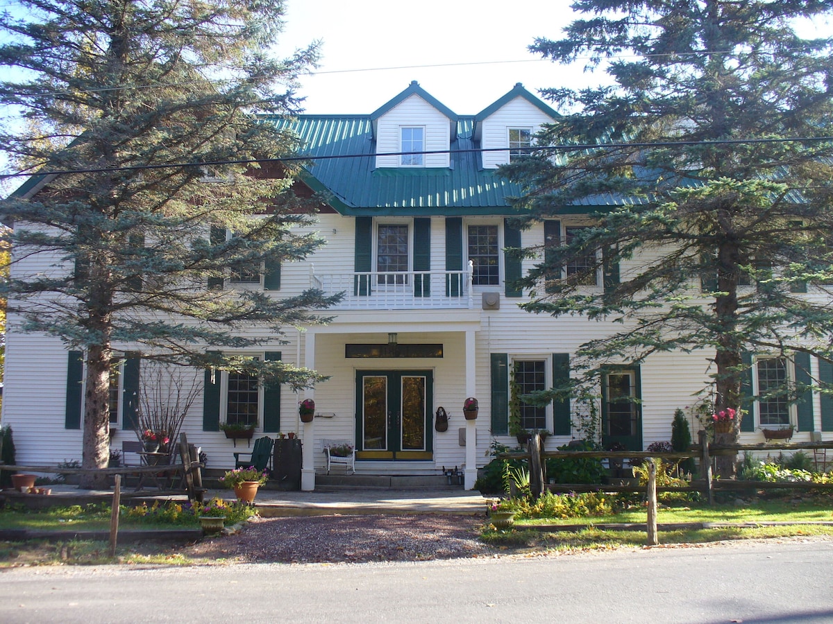 The exterior of a historic inn is displayed, featuring a white façade with green-shuttered windows and a prominent green roof. A spacious porch is visible, surrounded by lush gardens and tall trees that create a welcoming entrance.