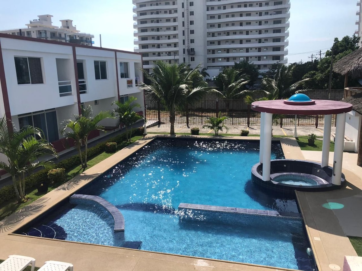 A spacious outdoor pool area features clear blue water and a circular gazebo nearby. Lush palm trees are positioned around the pool, and modern buildings provide a backdrop. Sun loungers are available on the side, inviting relaxation in the sun.