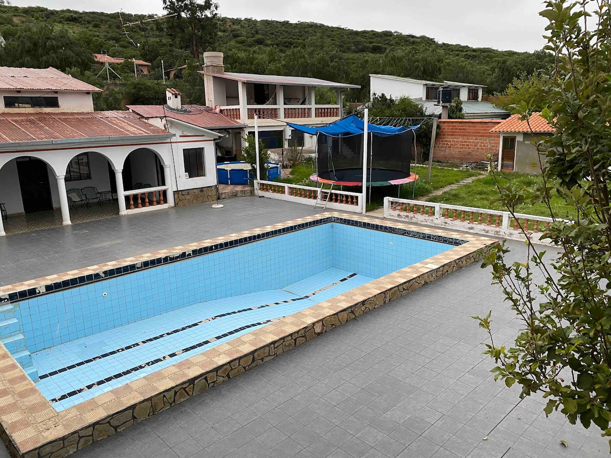 An outdoor area displays a rectangular swimming pool with light blue tiles, surrounded by gray tile flooring. Nearby, a trampoline is set up on green grass, with a trampoline frame visible. Multiple buildings with stucco walls and roofs can be seen in the background.
