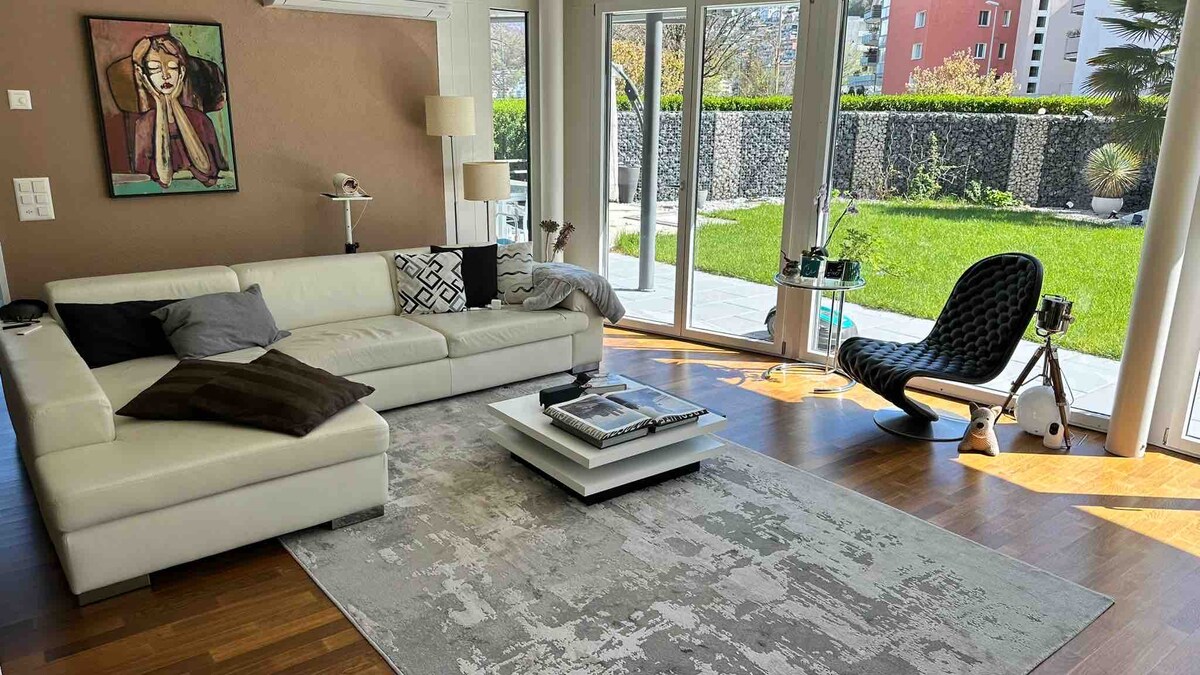A modern living area features a white sectional sofa positioned on a light-colored rug. Natural light floods the space through large glass doors that offer a view of the green garden outside. A sleek chair and coffee table complete the inviting seating arrangement.