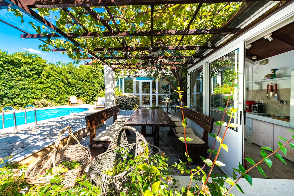 An outdoor area features a large dining table shaded by a trellis, surrounded by greenery. Woven chairs are positioned nearby. A swimming pool is visible in the background, with a small outdoor kitchen adjacent to the dining space, inviting relaxed gatherings.