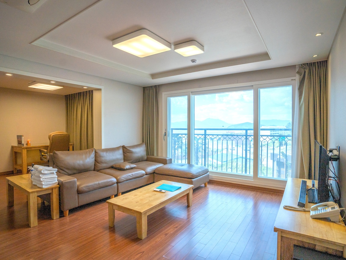 A spacious living area features a large sectional sofa arranged around a wooden coffee table. Bright natural light floods the room through a set of large windows, offering views of the surrounding landscape. The décor is neutral, creating a calm and comfortable atmosphere.